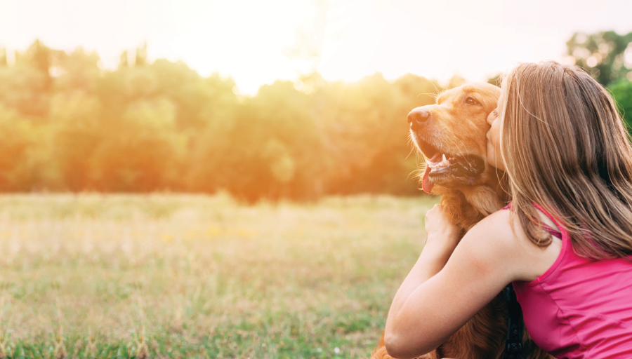 Kvinna sitter med sin hund utomhus på ett fält, kyssandes hunden.